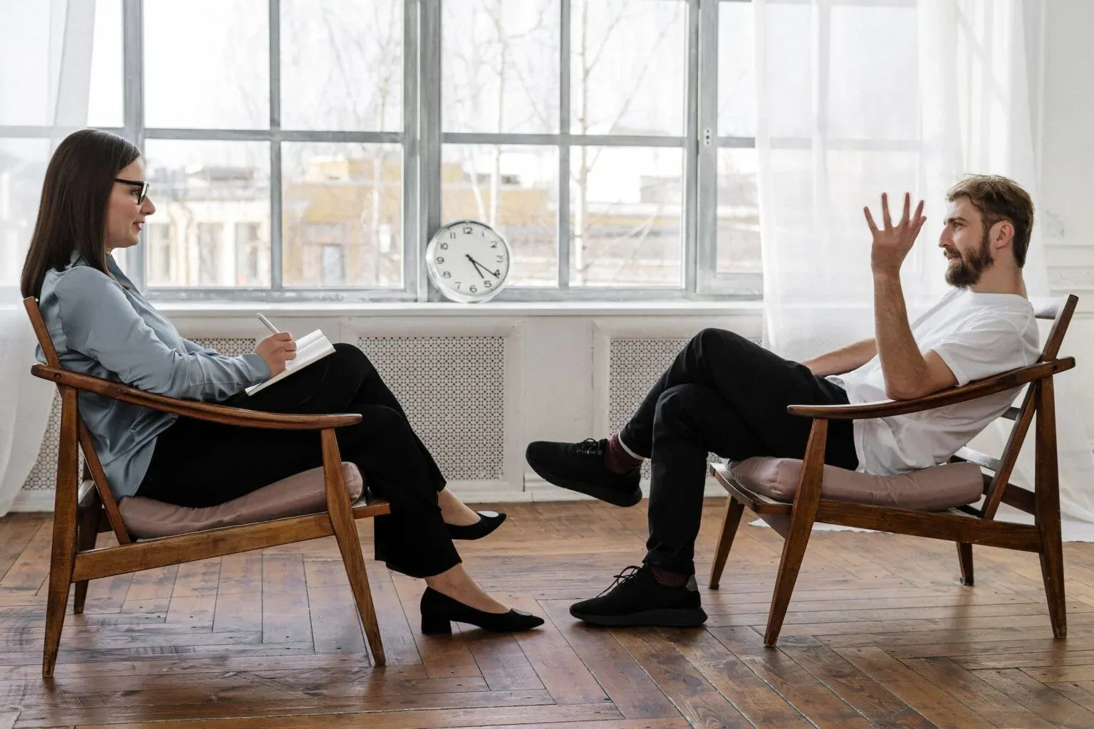 A therapist is sitting in an armchair, taking notes, while a man is talking to her while seated in another chair.