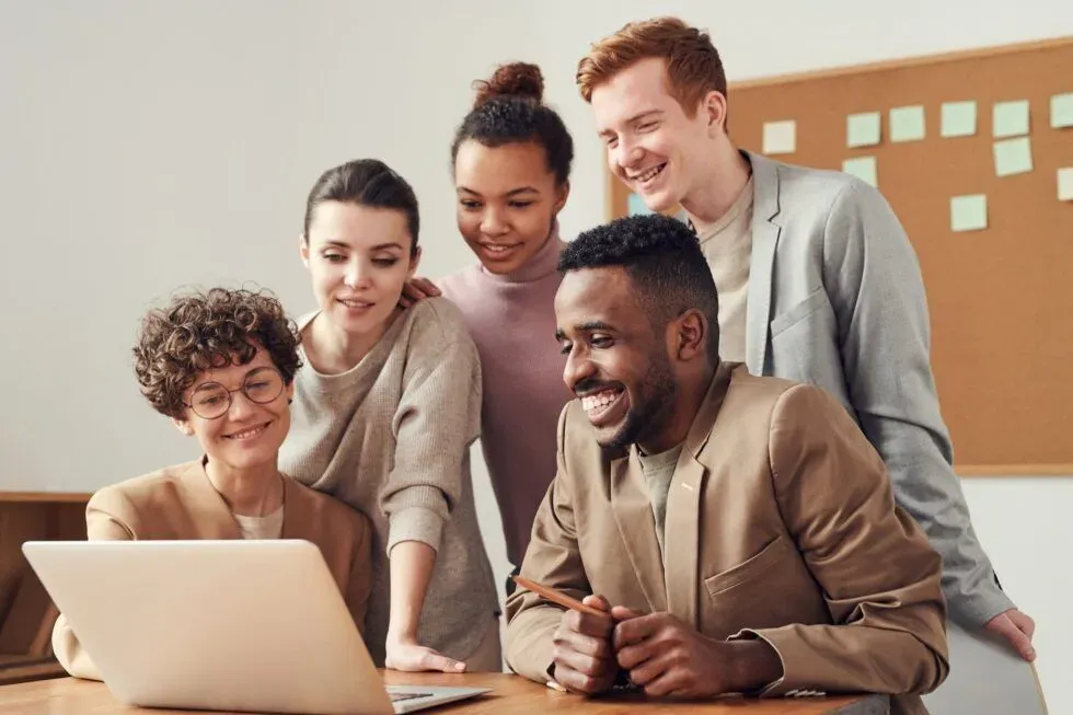 Five diverse young adults gathered around a laptop, engaging with the screen and smiling.