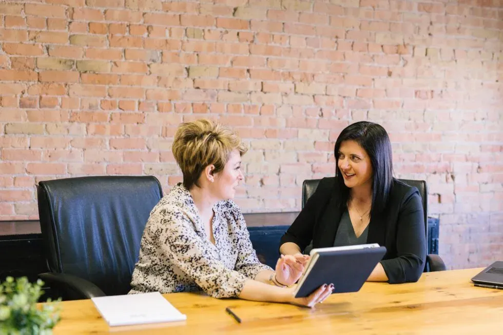 Two women sitting at a table with a tablet, engaged in conversation in a modern office setting with a brick wall.