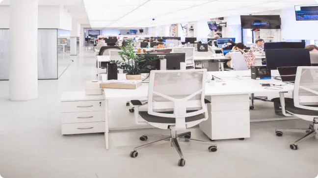 An open-plan office space with white desks, ergonomic chairs, and modern furnishings. Several employees are working at their desks with computers and office supplies. The environment is bright and organized, featuring large windows and a clean aesthetic.