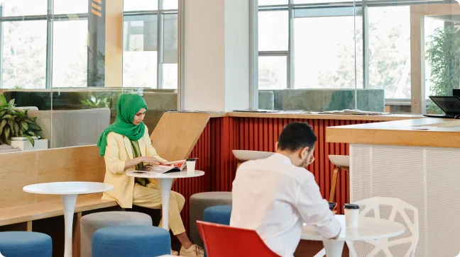 A woman wearing a green hijab sits at a table reading a magazine, while a man in a white shirt sits nearby, focused on his device. The setting appears to be a modern cafe or lounge with light-colored furnishings.