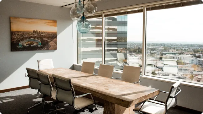 A modern conference room featuring a large wooden table and several white chairs, with a landscape artwork on the wall and large windows showcasing a city view.