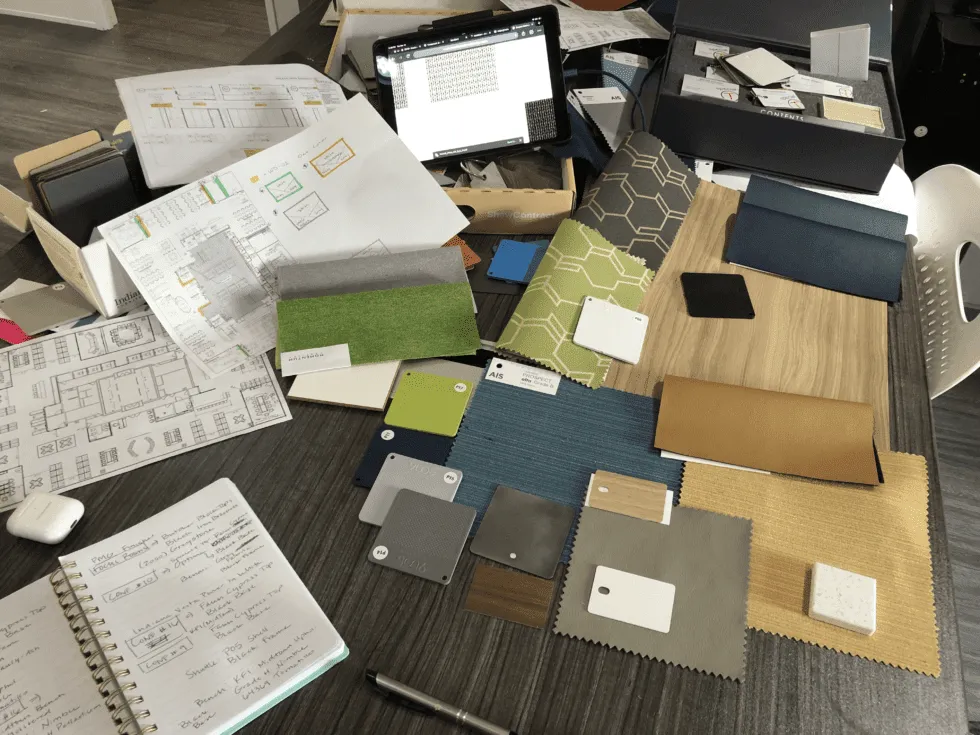 A cluttered workspace featuring various color samples, fabrics, papers, and design plans scattered across a table.