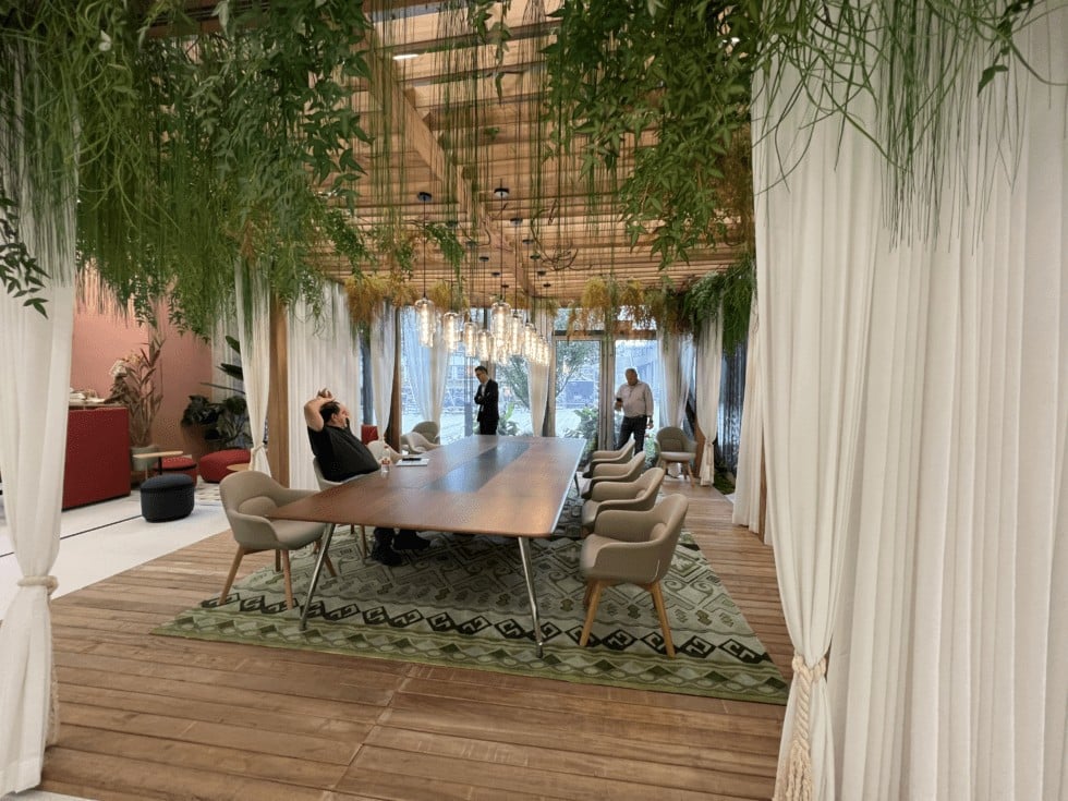 A modern meeting room with a large wooden table surrounded by chairs, greenery overhead, and sheer curtains, featuring a wooden floor.