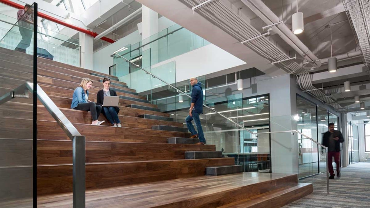 A modern office interior featuring a staircase with wooden steps, where two people are sitting with a laptop, while another individual walks up the stairs.