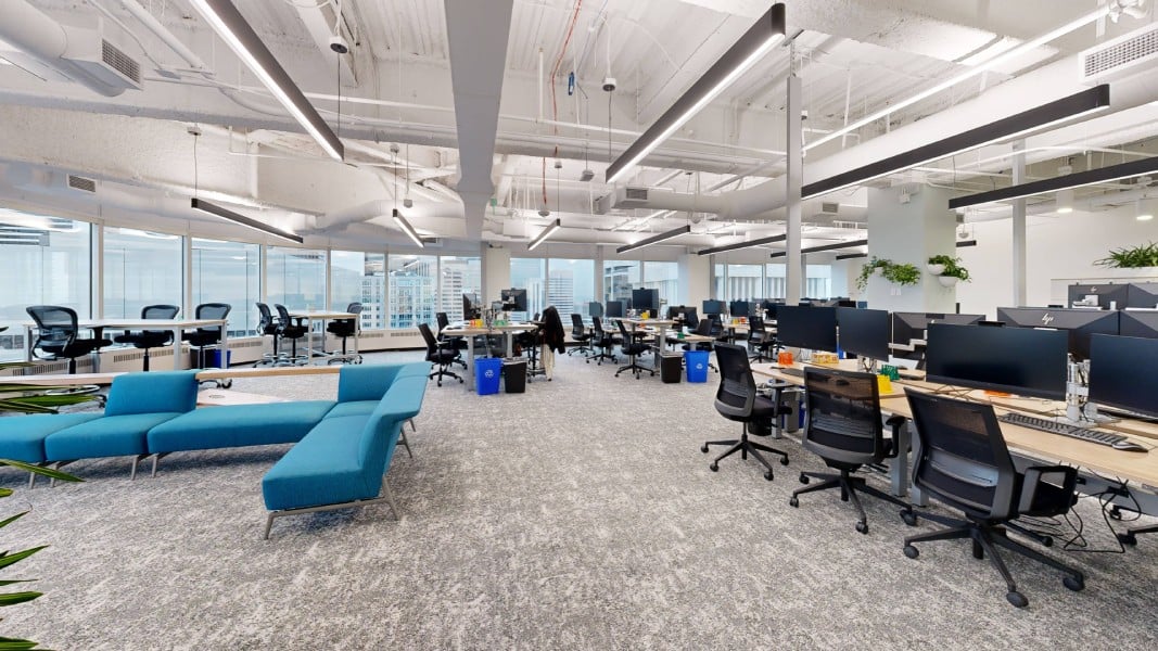 An open office space with modern furniture, including a blue couch and several workstations equipped with computers, surrounded by large windows.