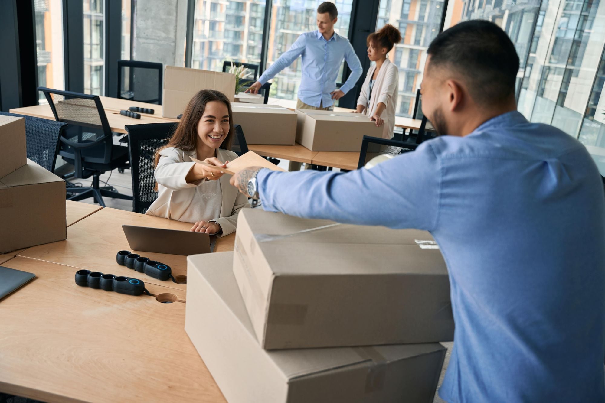 A group of professionals engaging in work in an office environment, with one person handing a document to another, while two others are seen in the background. Boxes are present on the table.