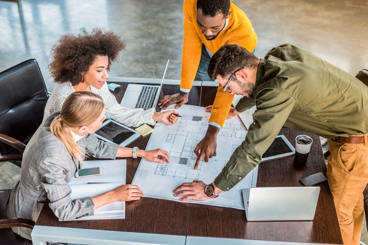 A group of diverse professionals collaborating around a table with blueprints and laptops.