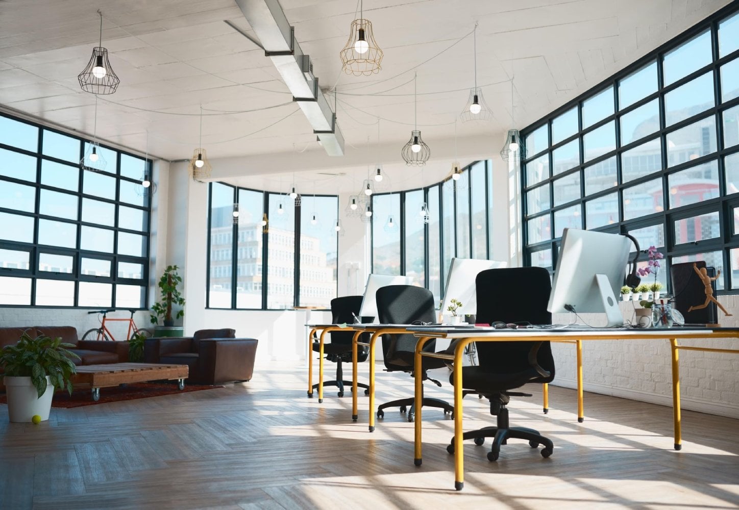 A modern office space featuring large windows, several desks with office chairs, decorative plants, and a comfortable seating area.