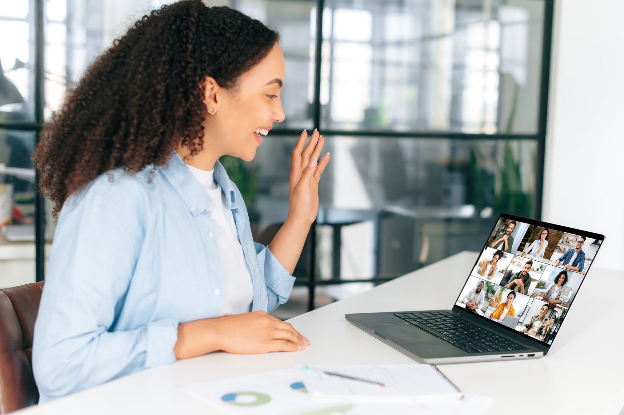 A woman with curly hair waves at her laptop while participating in a video call, showing a grid of people on the screen. She is seated at a desk with some papers and office supplies around her.