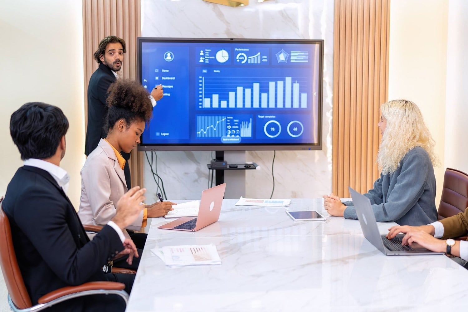 A diverse group of professionals in a boardroom meeting, one person presenting data on a large screen, while others are seated at a conference table with laptops.