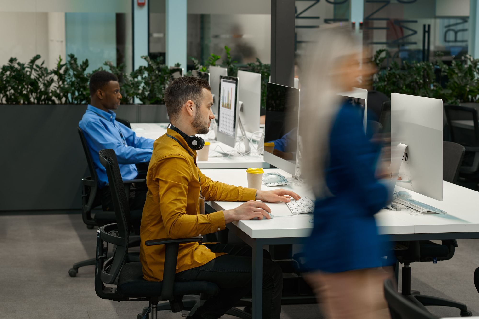 A modern office space with three people working at desks. One person is focused on their computer, wearing headphones and a mustard-colored shirt. Another person is seated in the background, wearing a blue shirt. A blurred figure in a blue outfit is walking past.