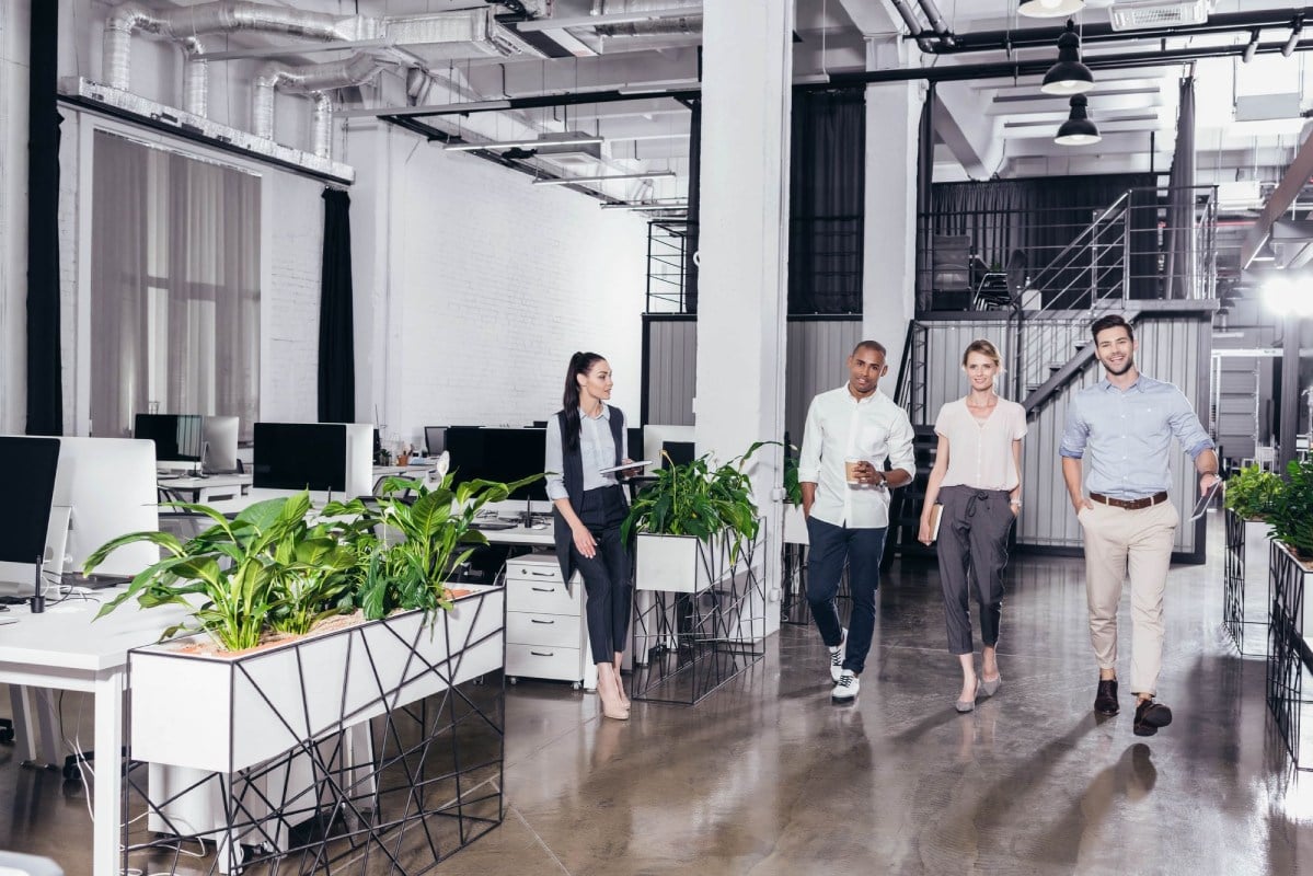 A group of four professionals walking through a modern office space with greenery and workstations.