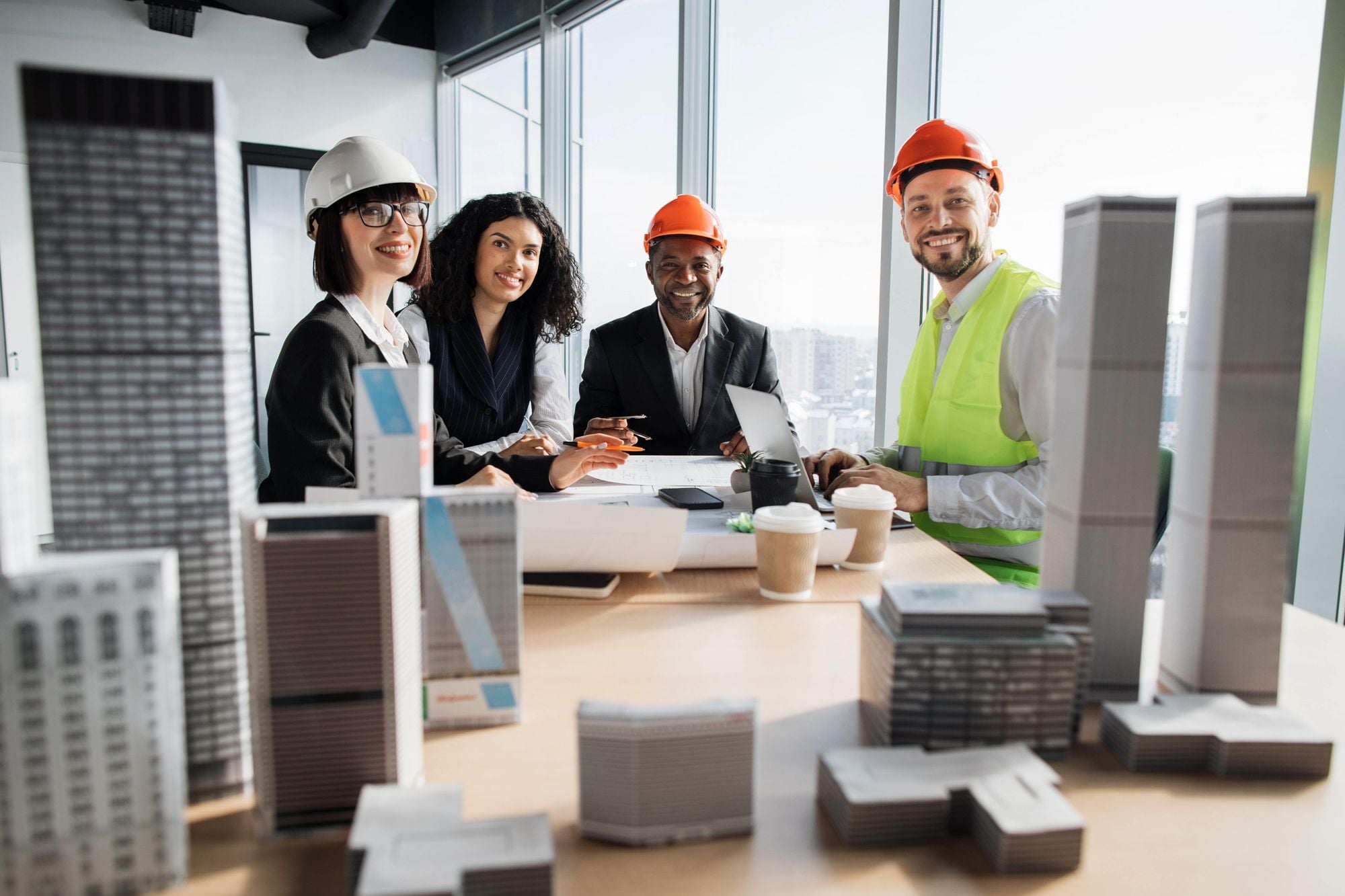 A group of four professionals, including two women and two men, are gathered around a table with architectural models and documents. They are discussing ideas, wearing hard hats and safety vests, with a city skyline visible through the windows behind them.