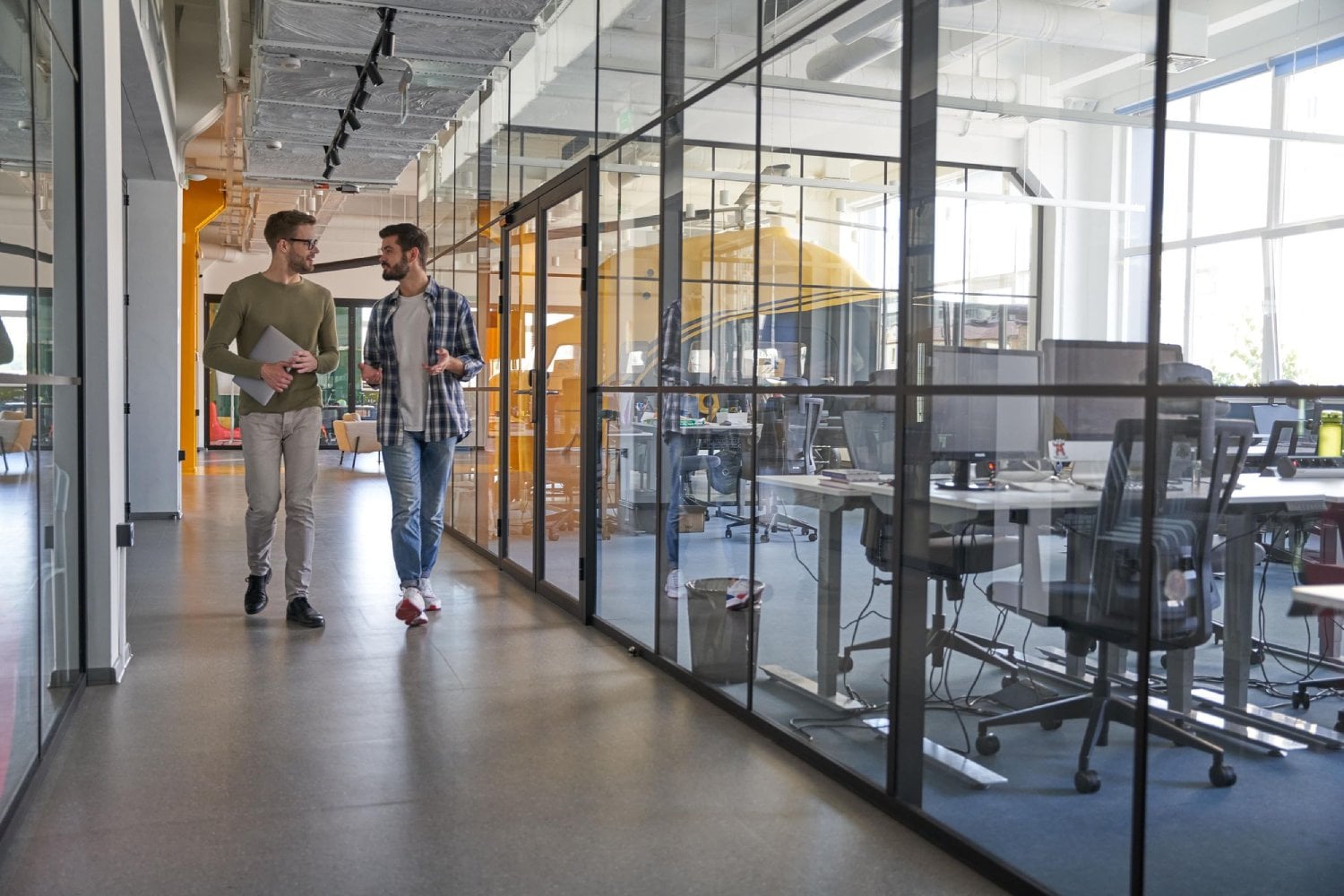 Two men walking and talking in a modern office space with glass partitions and empty workstations.