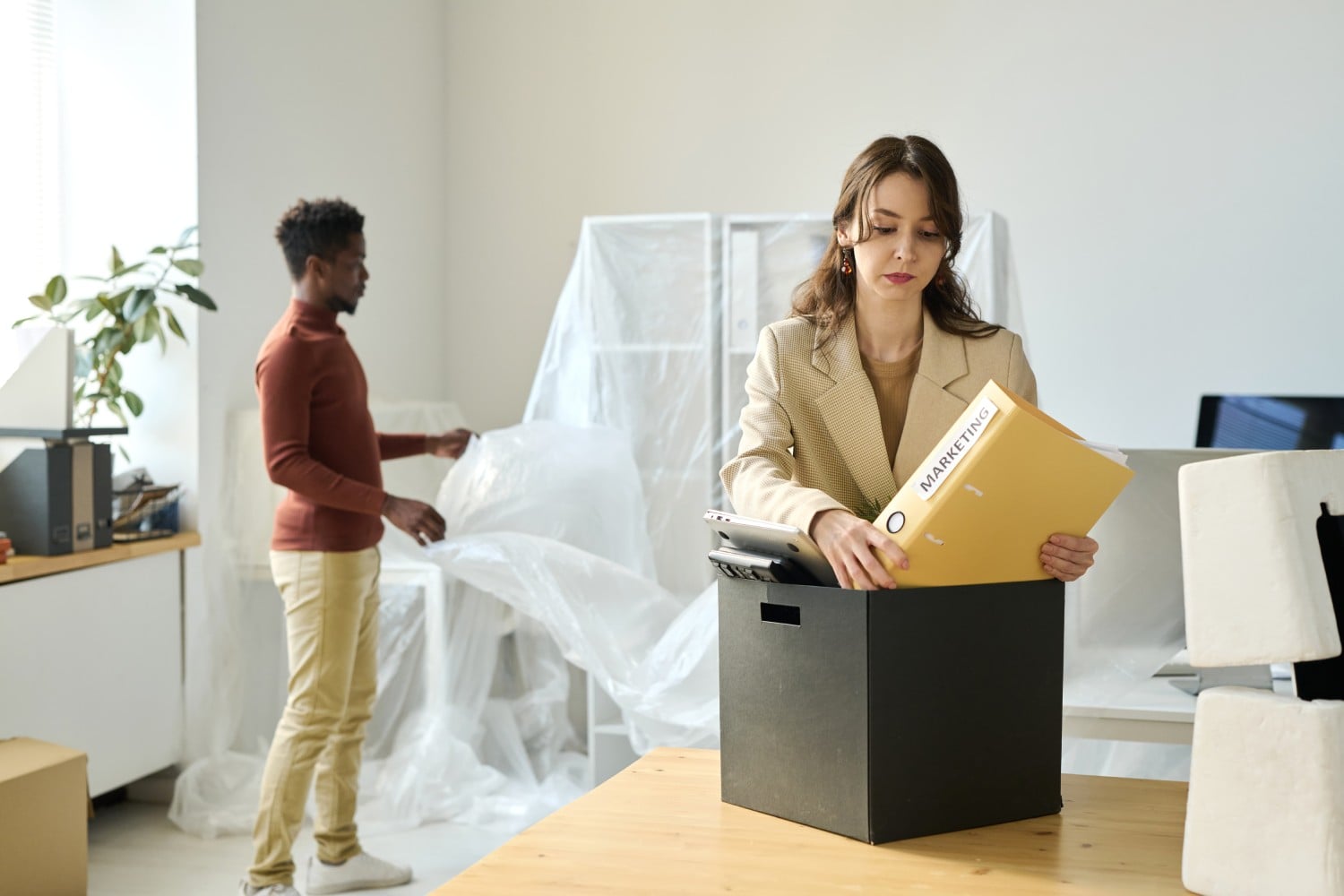 A woman in a blazer places files into a black box while a man in a sweater looks at a transparent sheet in an office setting.