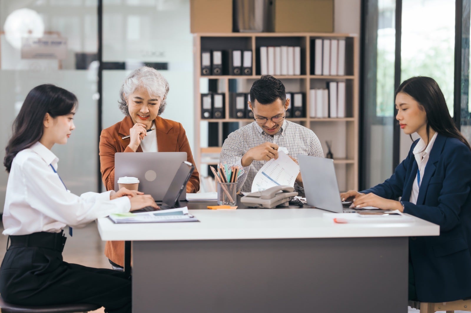 Four people working collaboratively at a modern office table, using laptops and other devices, with office supplies around them.