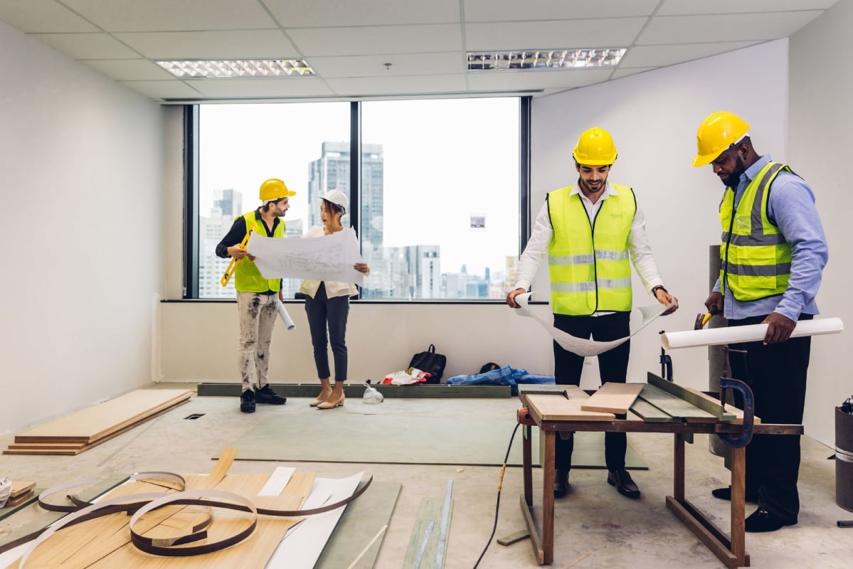 Four construction workers in hard hats and safety vests are inside an unfinished office space, reviewing plans and working with materials.