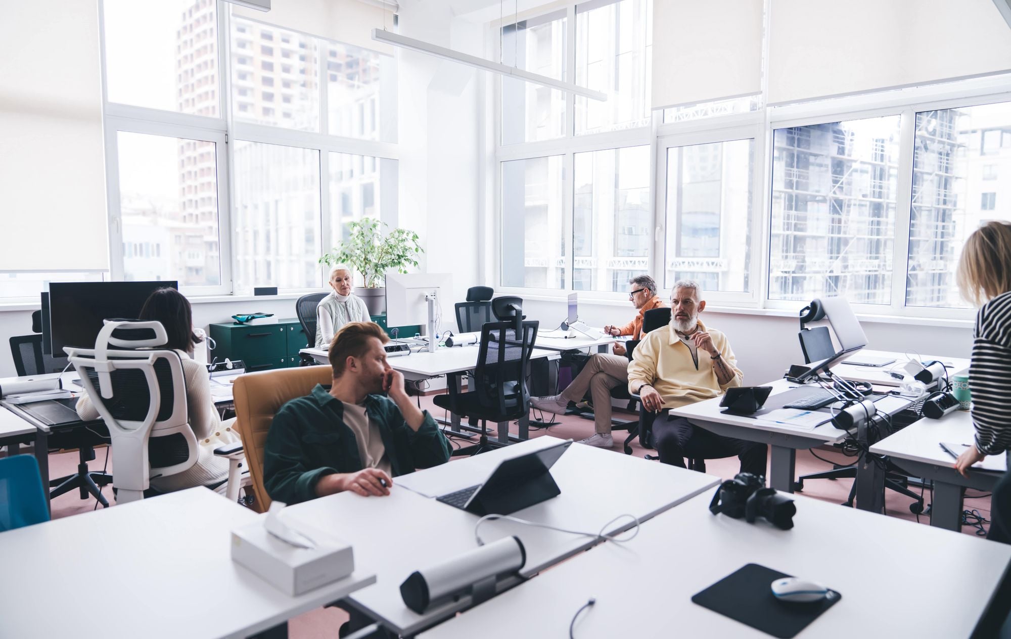 A modern office space with several people engaged in discussion, surrounded by desks, computers, and large windows letting in natural light.