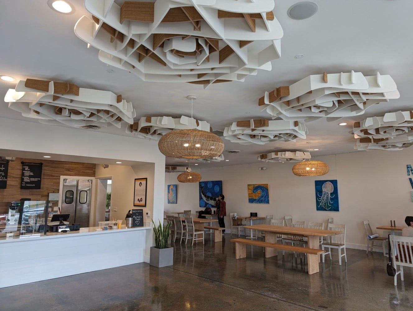 A modern café interior featuring unique ceiling designs, a counter with a display case, multiple artworks on the walls, and various seating arrangements.