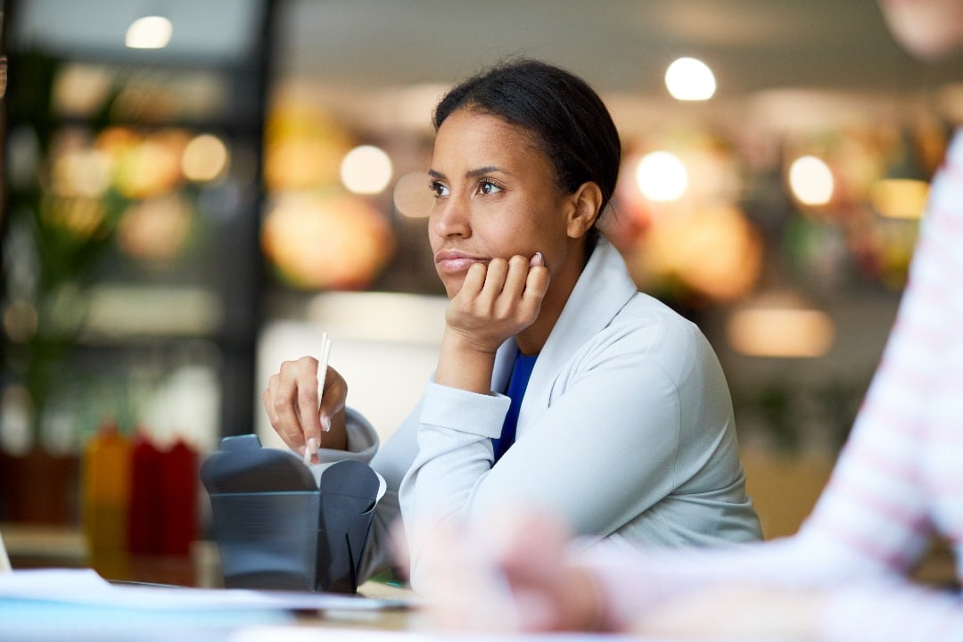 A woman sitting at a table with a thoughtful expression, resting her chin on her hand, with a blurred background.