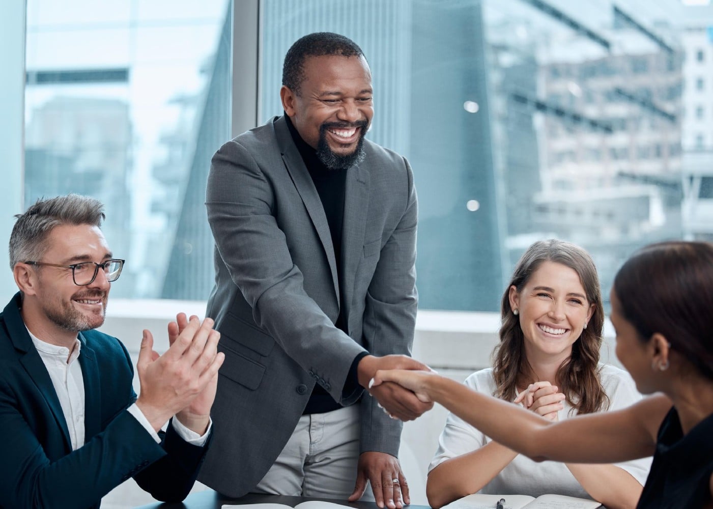 A group of professionals in a meeting, with a man shaking hands with a woman while others look on and smile.