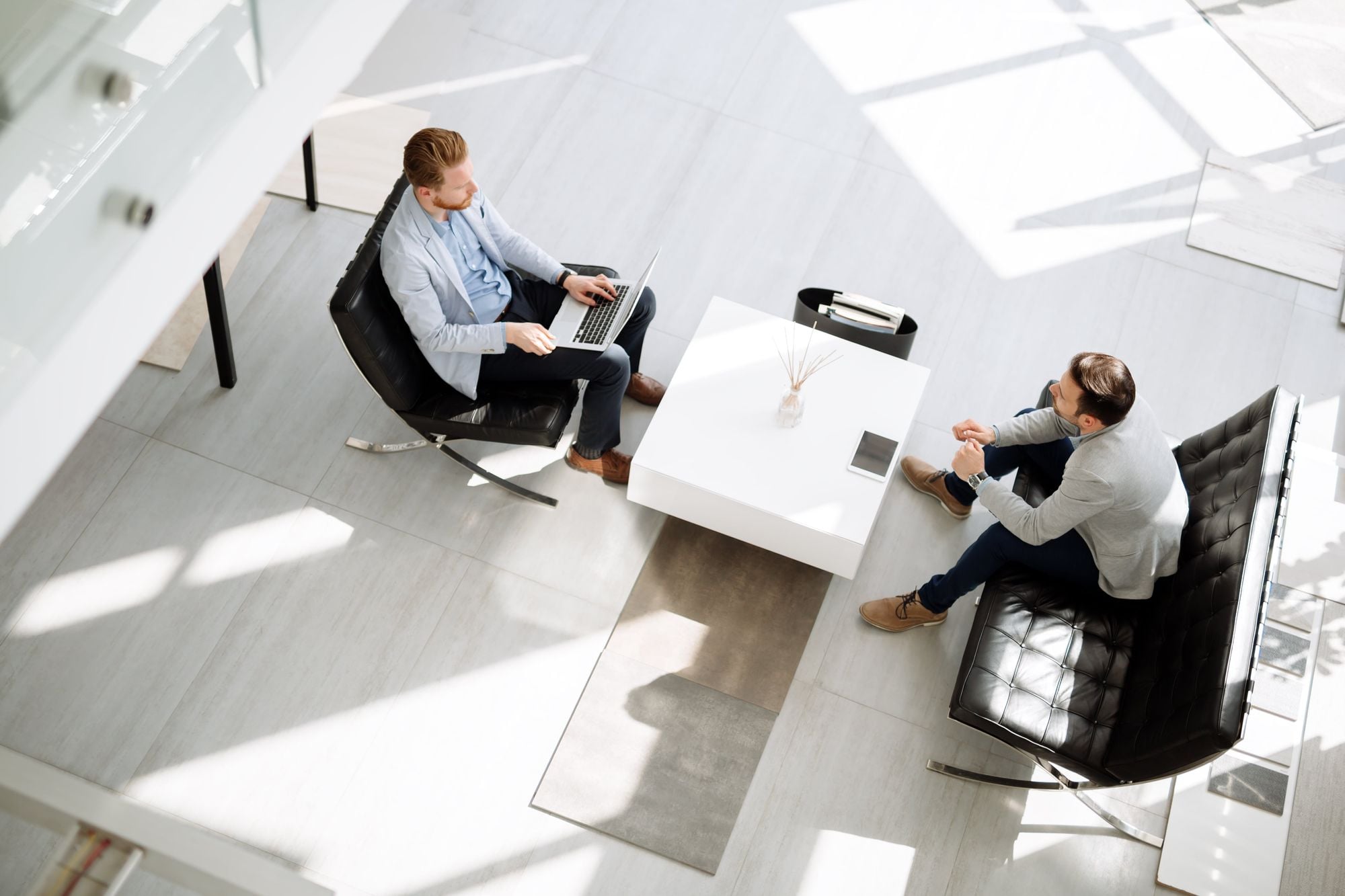 Two men sitting in a well-lit modern office space, one on a laptop and the other engaged in conversation, around a small white table.