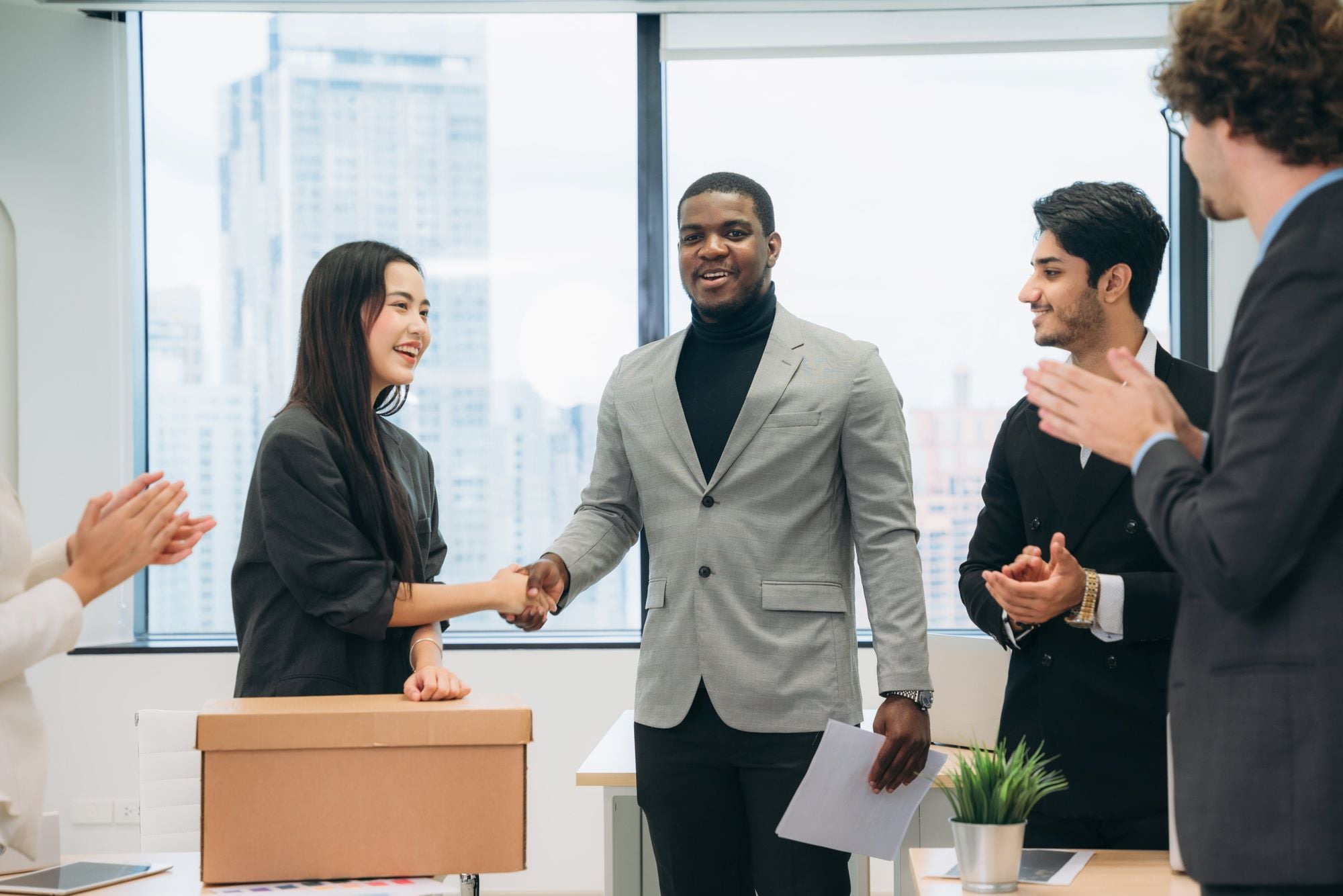 A group of professionals in a modern office celebrating a successful handshake between a woman and a man, with others applauding in the background.