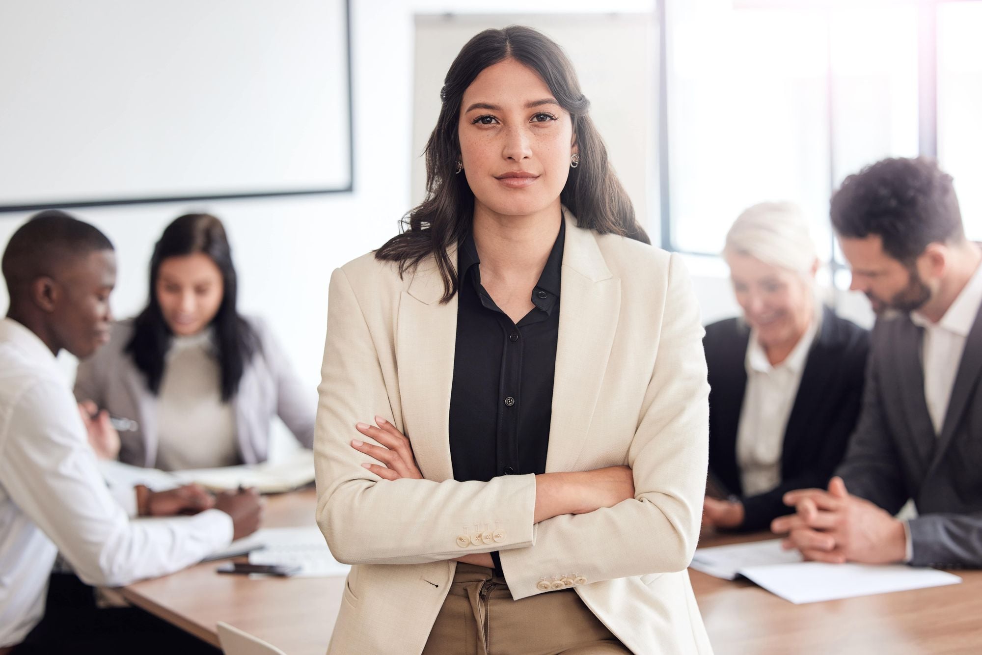 A confident woman in a blazer stands with arms crossed in a meeting room, while colleagues engage in discussion in the background.