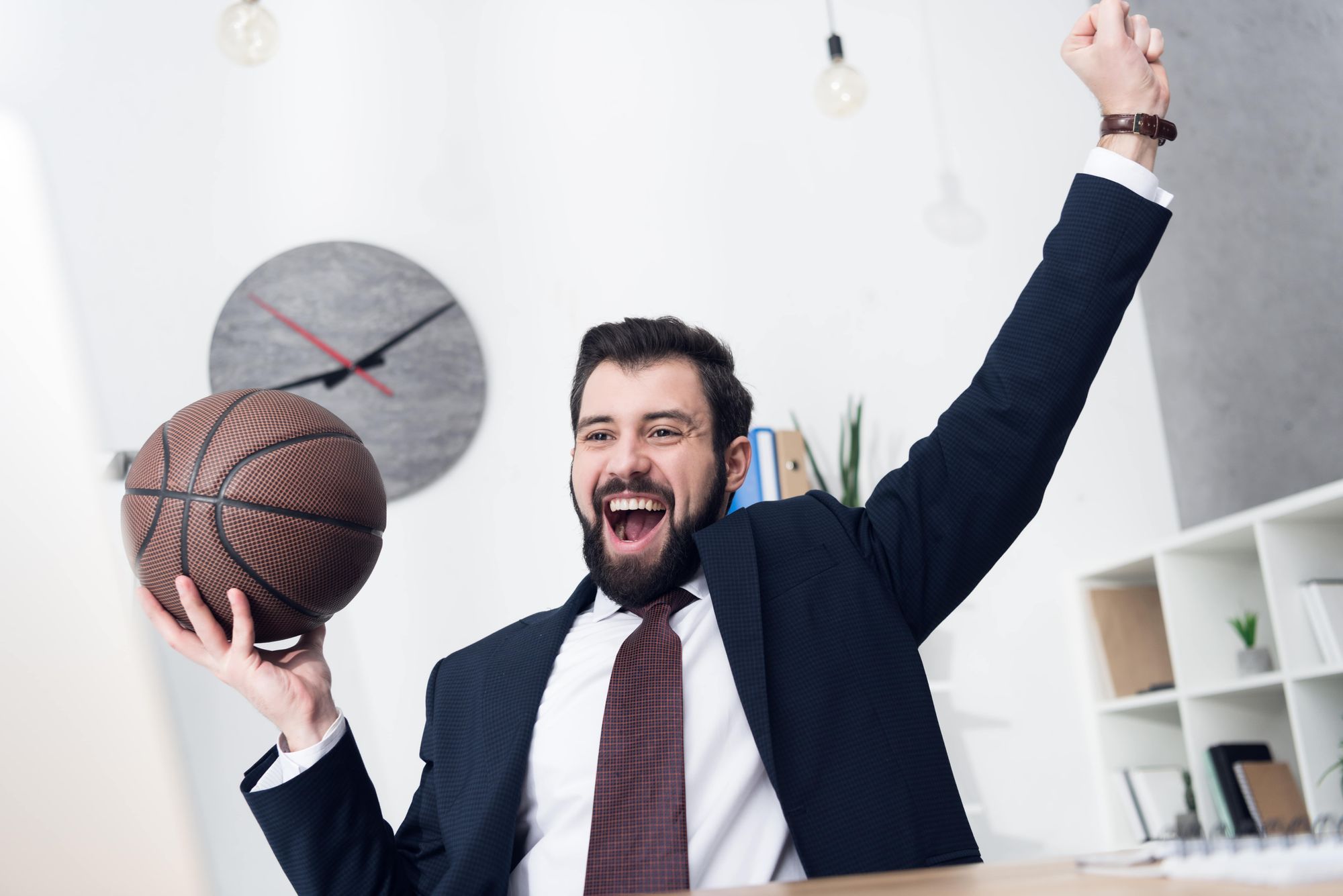 A man in a suit sitting at a desk, excitedly raising a basketball in one hand while celebrating.