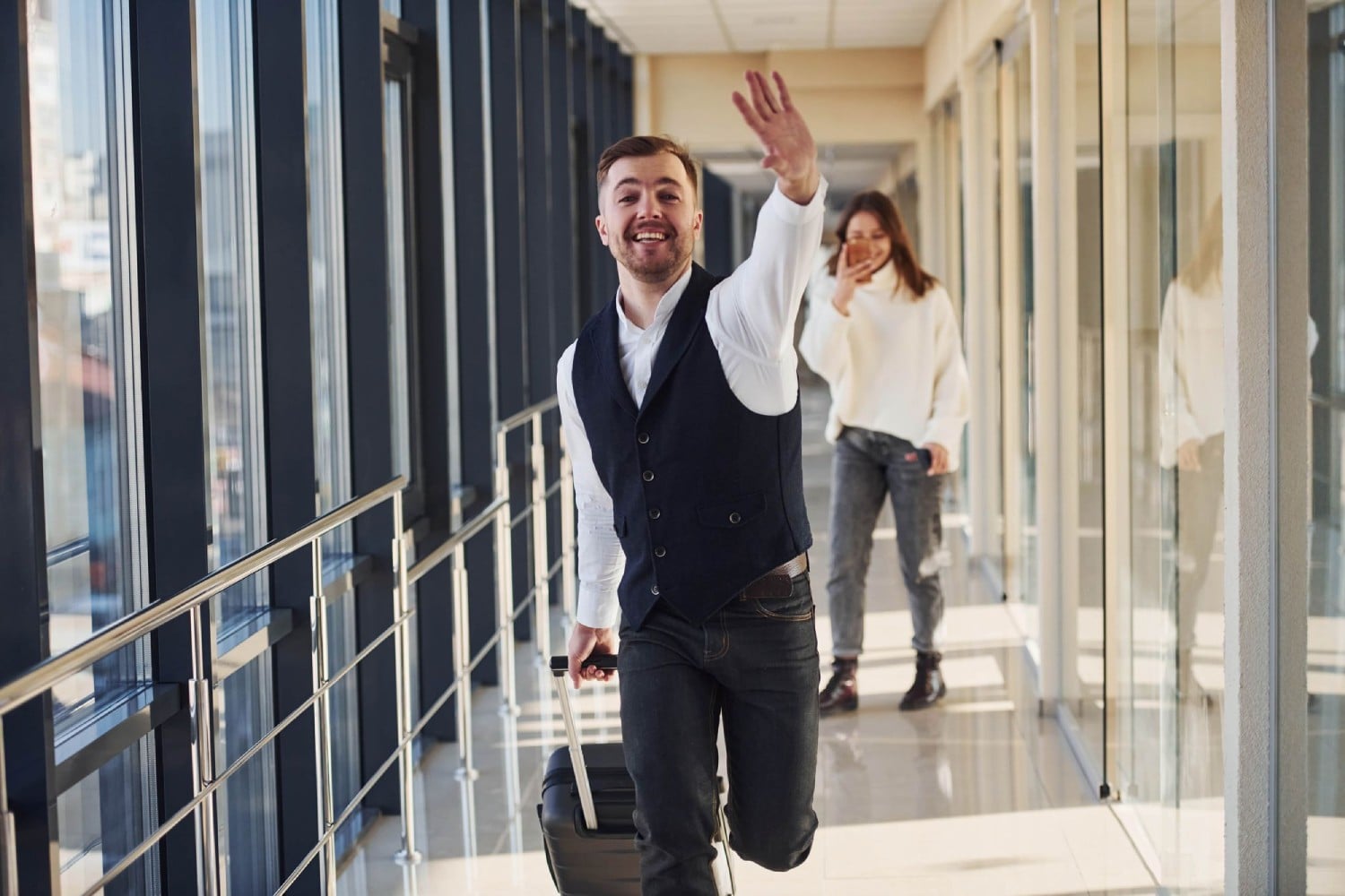 A man in a vest waves while walking down a corridor, pulling a suitcase, with a woman smiling in the background.