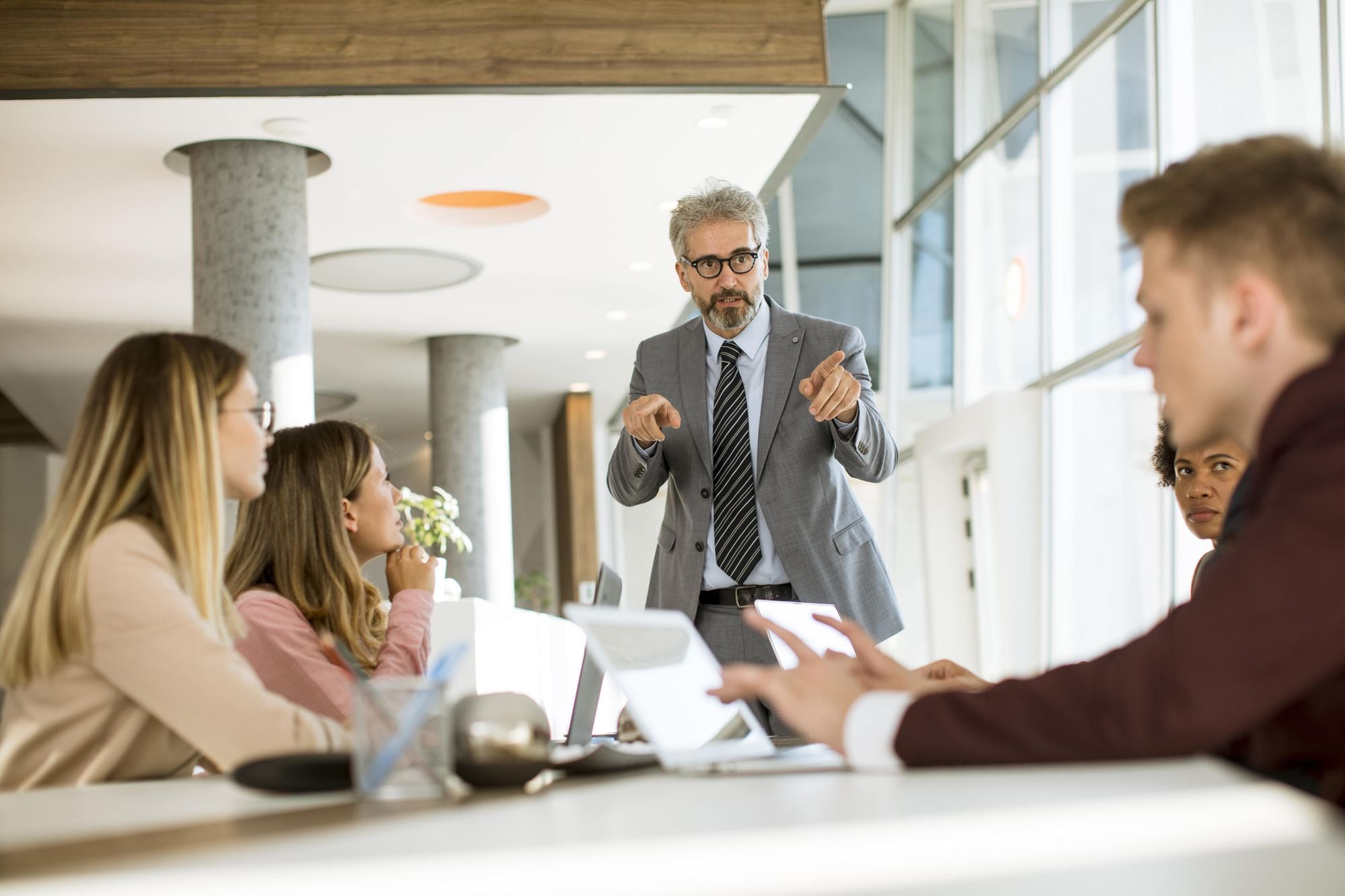 A group of professionals in a modern office setting, with a man in a suit leading a meeting while others listen attentively.