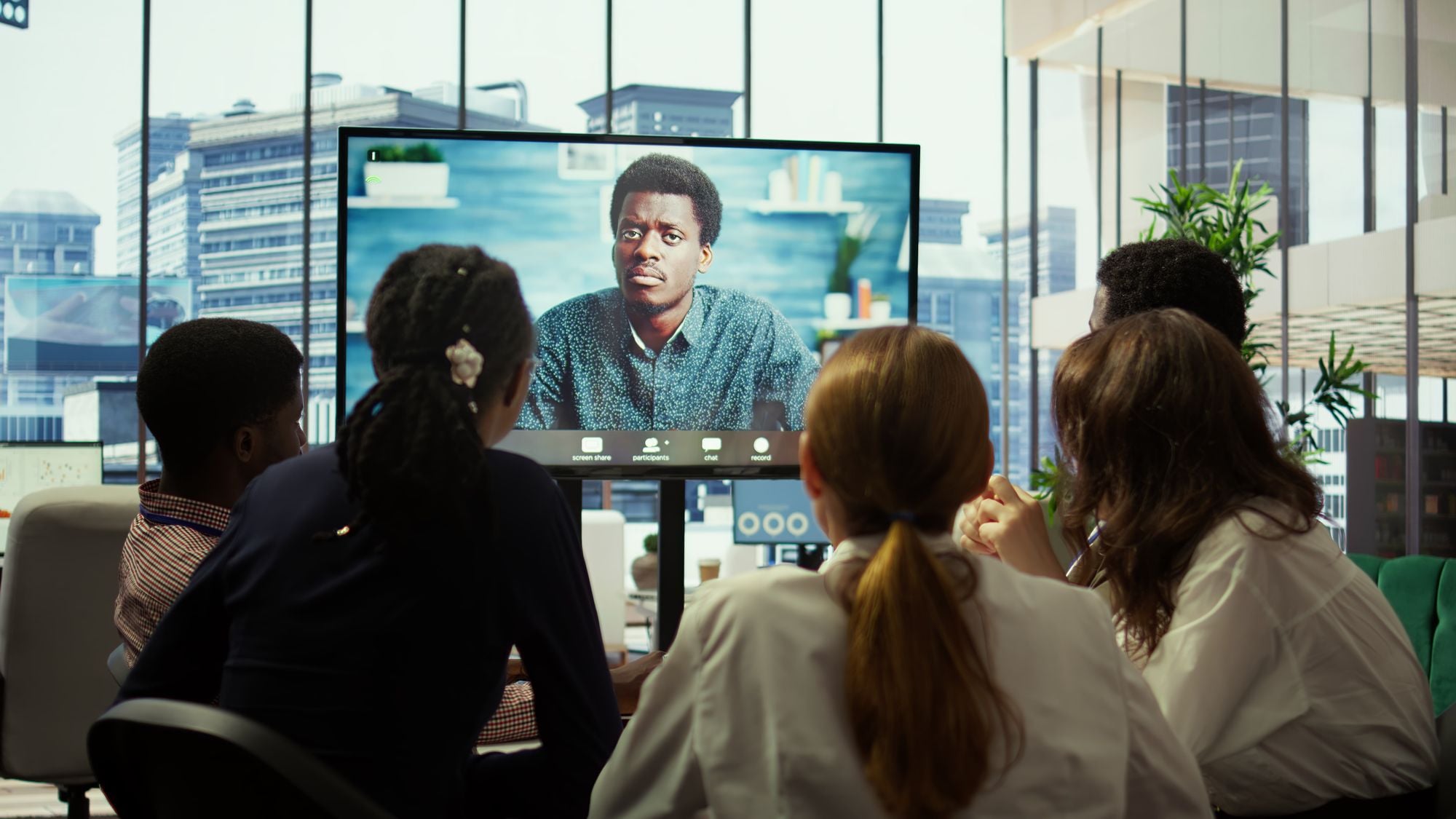 A group of people sitting at a table in an office, attentively watching a video call on a screen displaying a man.