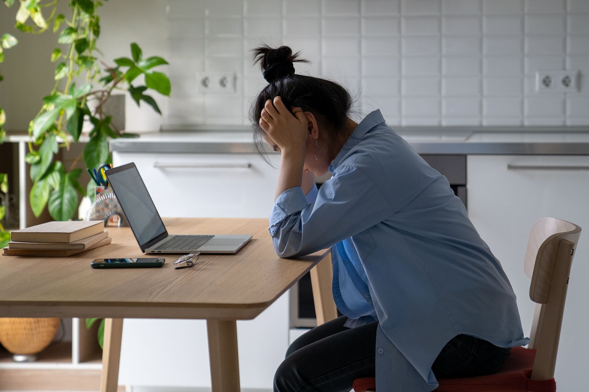 A person sitting at a table with a laptop, looking stressed or overwhelmed, with their hand on their head. There are plants and books in the background.