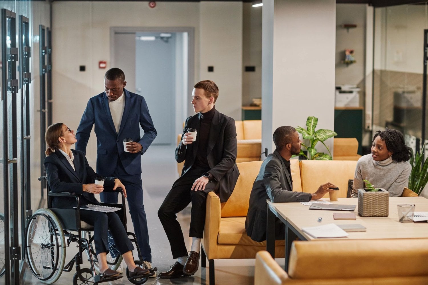 A diverse group of professionals engaging in conversation in a modern office setting. One person is in a wheelchair, while others are standing or sitting, holding cups.