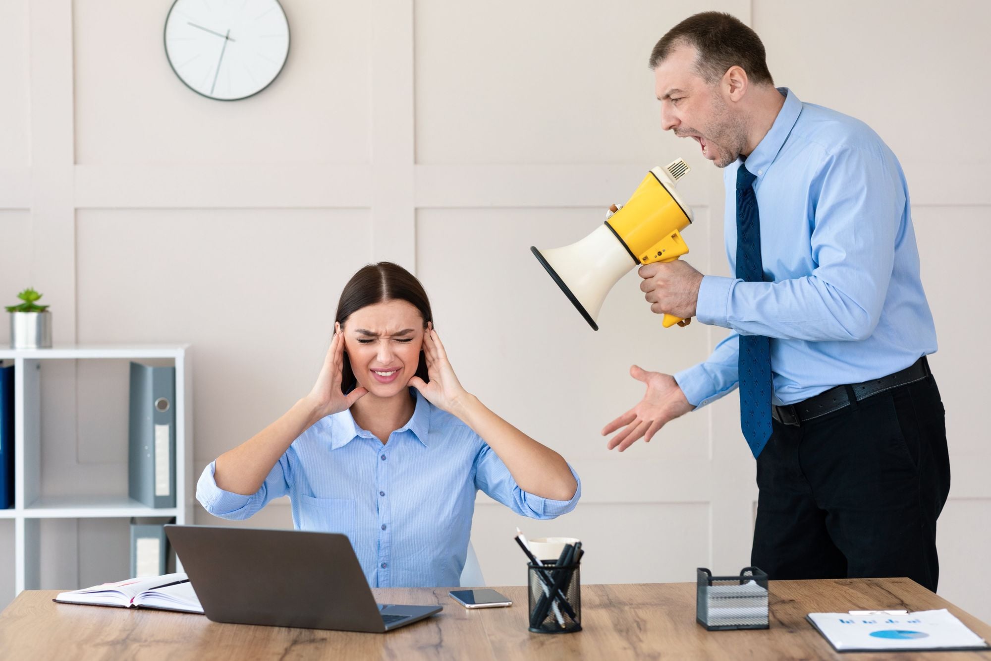 A man in a suit is shouting into a megaphone while a woman in a blue shirt is sitting at a desk, covering her ears in distress.
