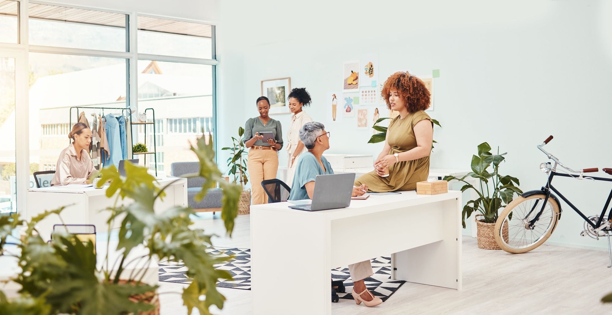 A bright and modern workspace with several people engaged in various activities; one person is sitting at a desk with a laptop, another is seated nearby, and several others are standing or interacting in the background with plants and a bicycle present.