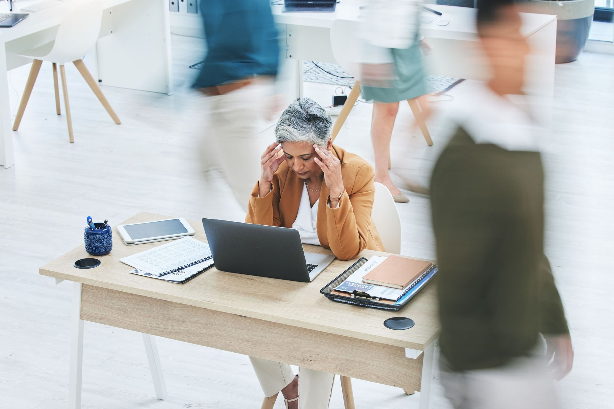 A woman sitting at a desk, looking stressed while working on a laptop, while people are walking by in a busy office environment.