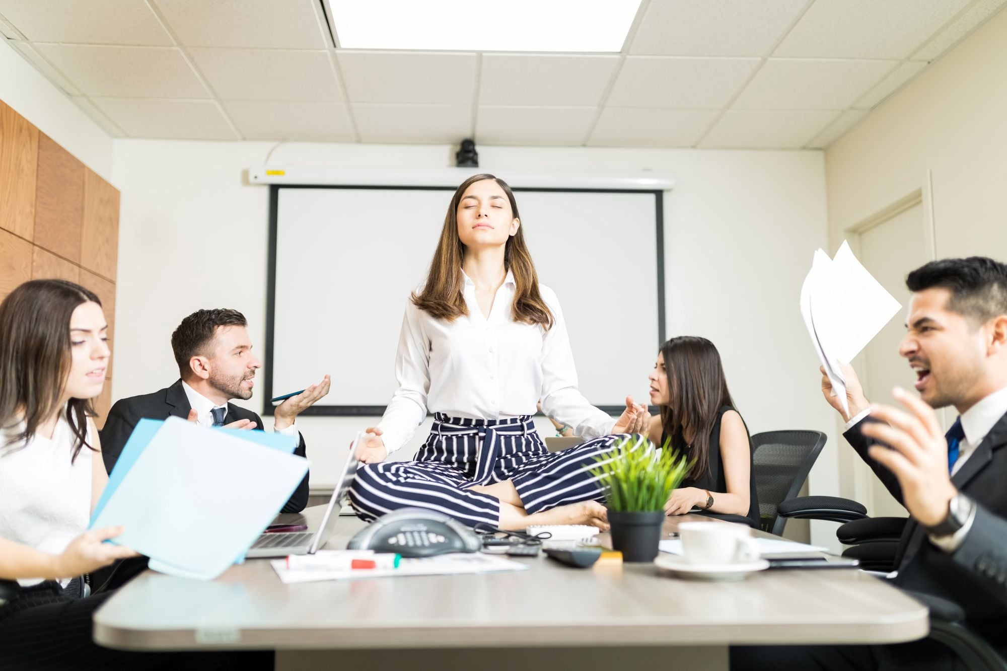 A woman sitting cross-legged on a conference table, meditating, while four colleagues around her appear to be arguing or stressed in a chaotic office meeting.