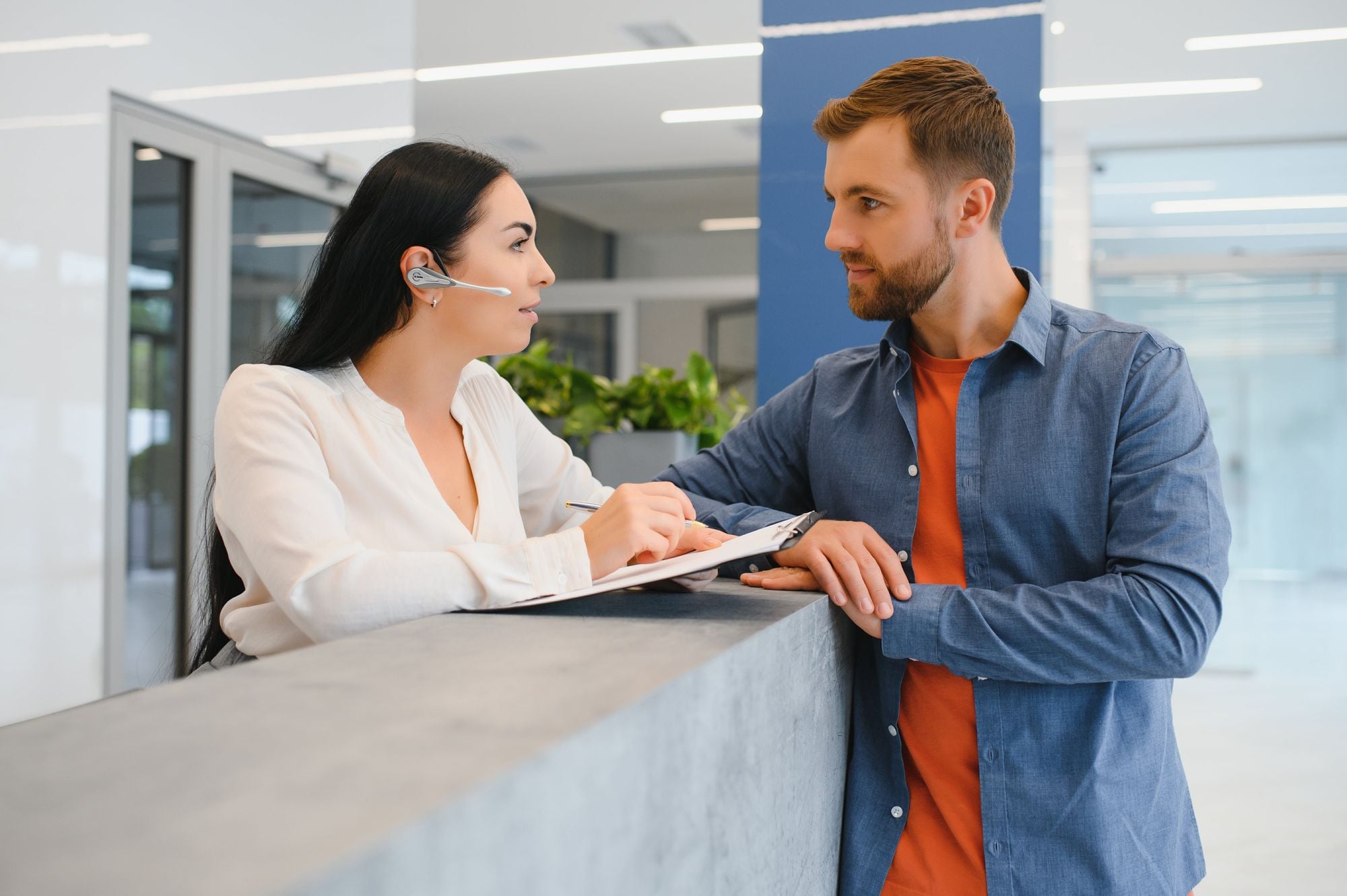 A woman with long dark hair wearing a headset and a white blouse is talking to a man in a blue shirt. They are both engaged in a conversation near a reception desk in a modern office setting.
