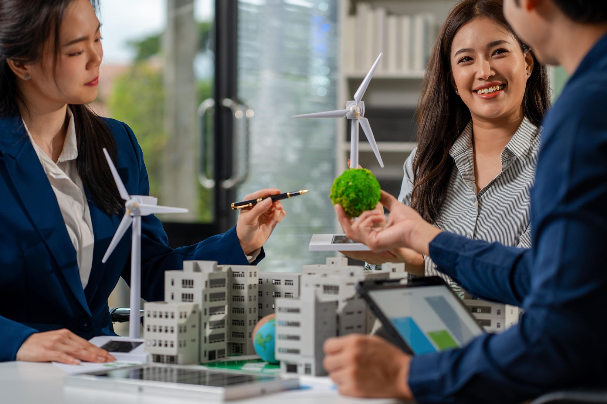 A group of professionals in a meeting, discussing sustainable architecture with models of buildings and eco-friendly elements like wind turbines and a green globe.