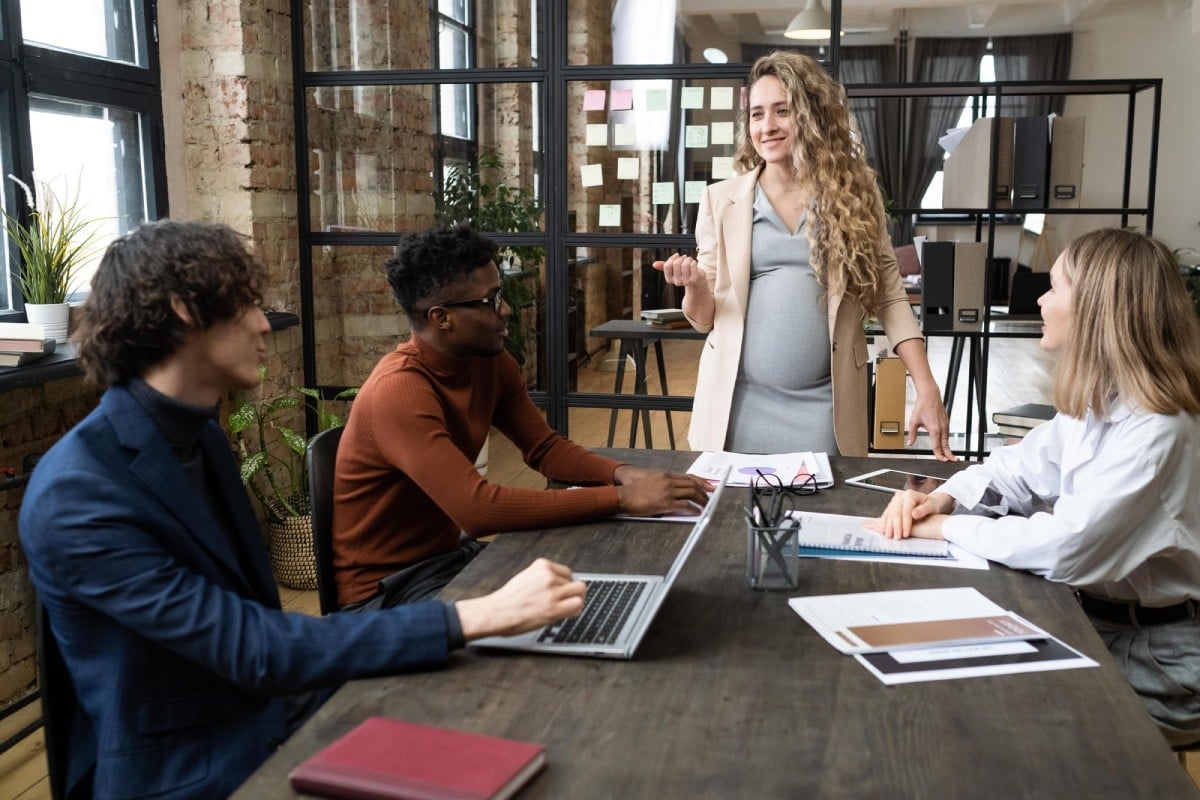 A group of four diverse individuals in a modern office setting, engaged in a discussion. One woman, who appears to be pregnant, is standing and speaking, while three others are seated at a table with laptops and documents in front of them. The space features large windows and industrial design elements.