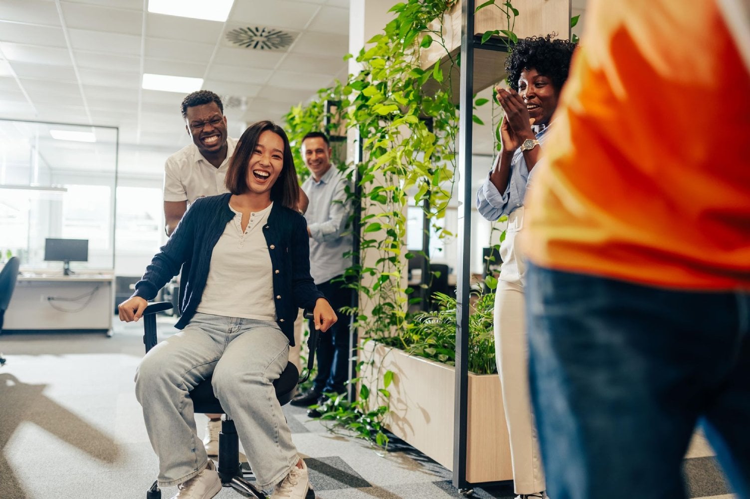 A group of four colleagues in an office are smiling and enjoying a moment, with one person sitting on a swivel chair being pushed by another. The atmosphere is cheerful and lively.