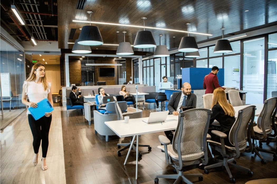 An open office space with modern furniture, where employees are working at desks and in casual seating areas. There are several people using laptops and discussing in groups, while a woman walks by holding a folder.