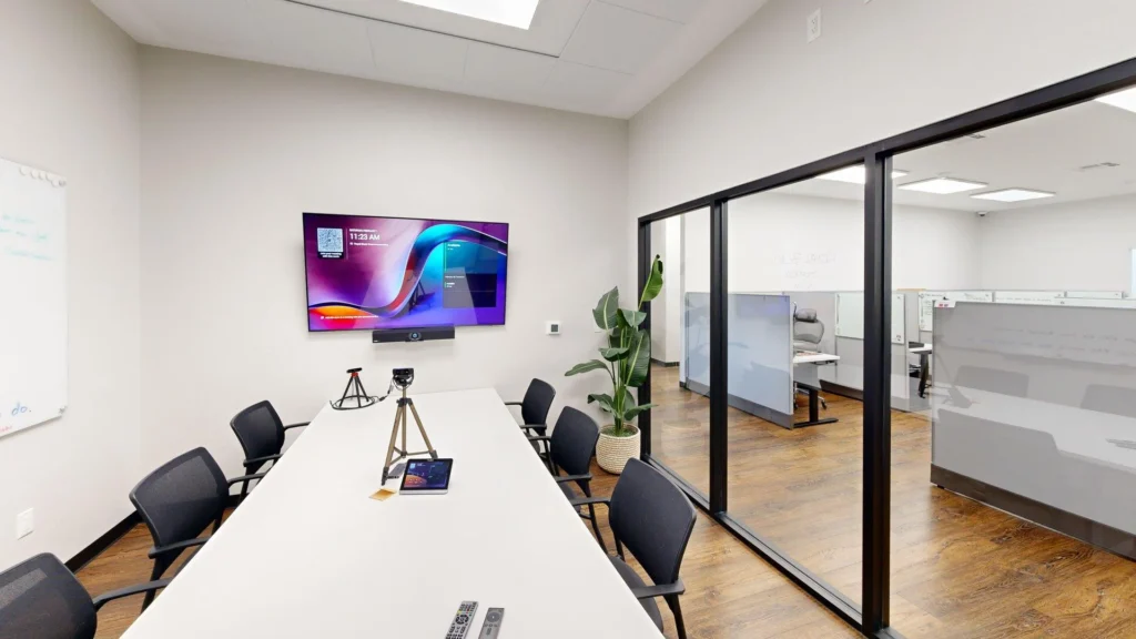 A bright meeting room featuring a long white table with black swivel chairs, a wall-mounted TV with AV equipment, a tripod setup, and floor-to-ceiling glass partition walls overlooking an office area.