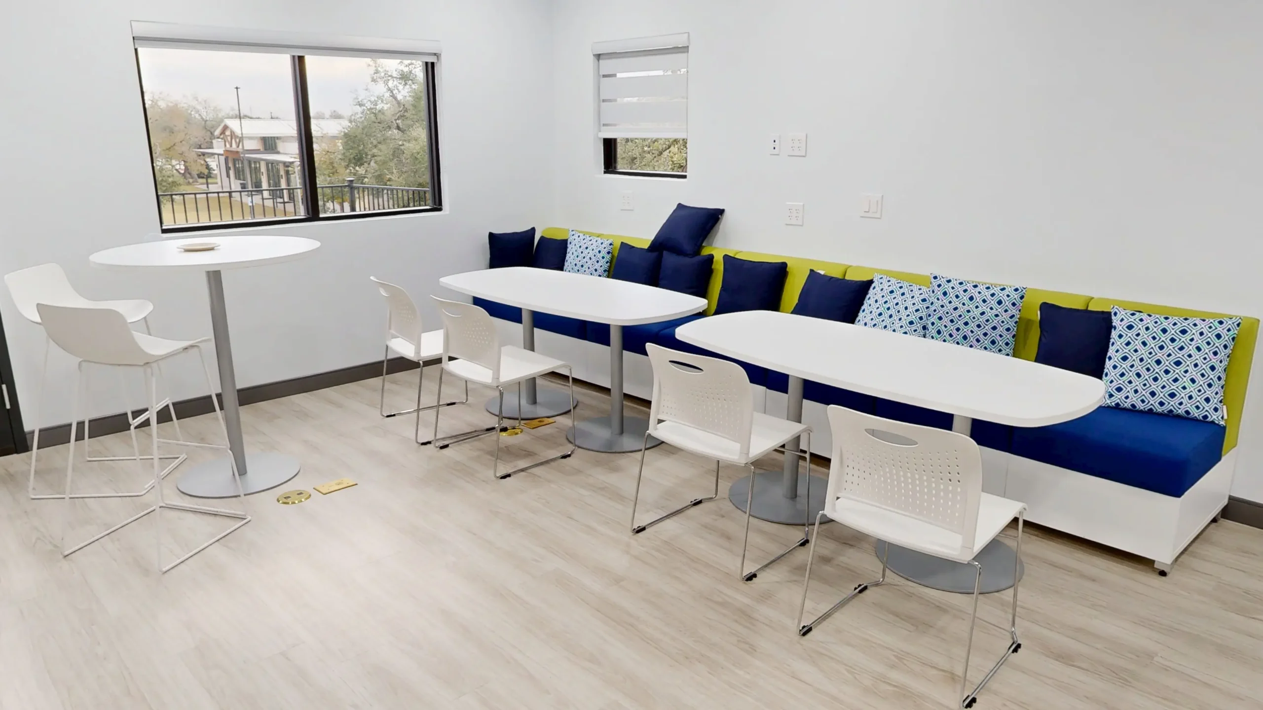 A modern office break room featuring light wood flooring, white dining tables with matching plastic chairs, and a long built-in banquette couch accented with blue and lime green cushions.