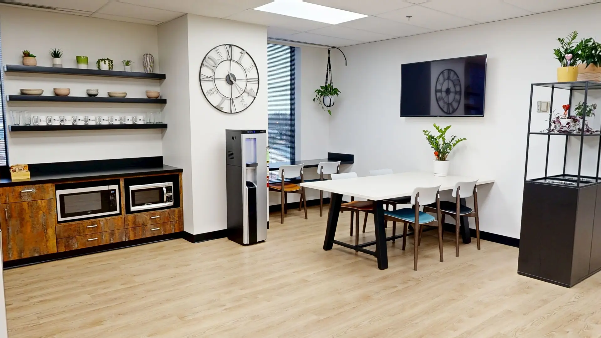 A modern office kitchenette and break area featuring floating shelves lined with coffee mugs, built-in microwaves, a water dispenser, a white dining table surrounded by colorful chairs, and a large metal wall clock.