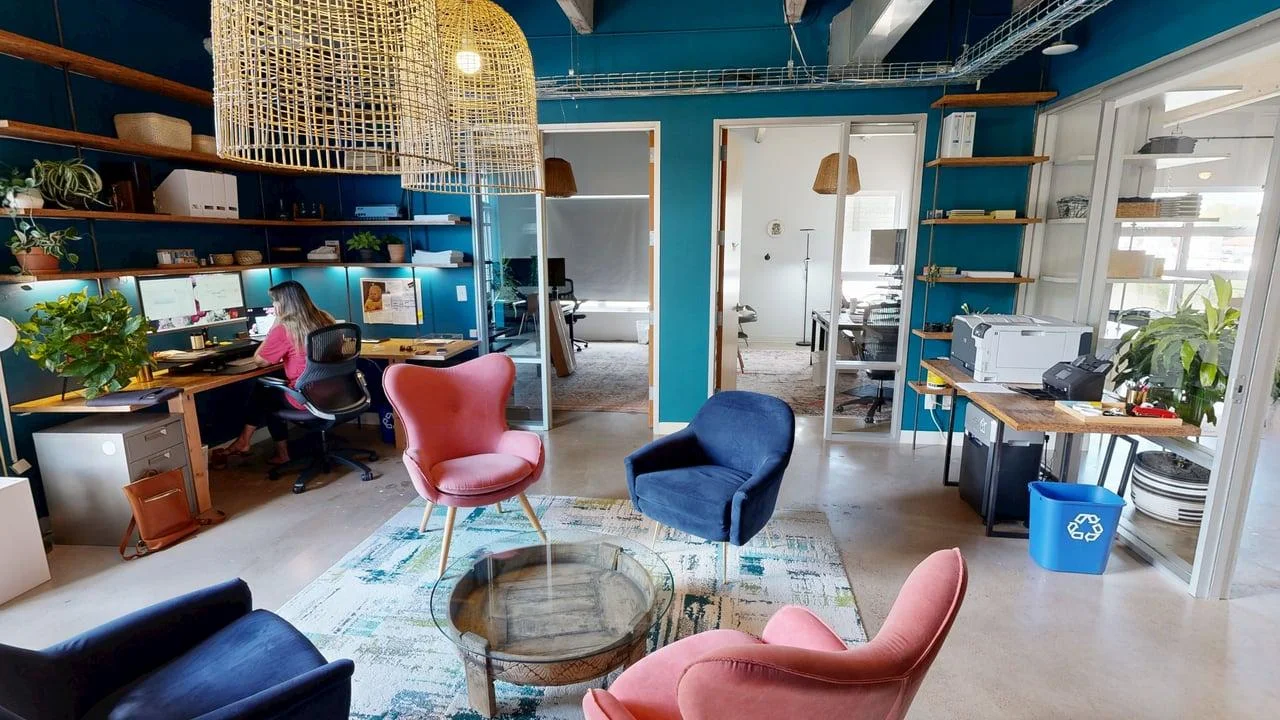 A vibrant office common area featuring teal walls, large woven basket pendant lights, a central seating lounge with pink and dark blue armchairs around a round coffee table, and an adjacent built-in desk workstation.