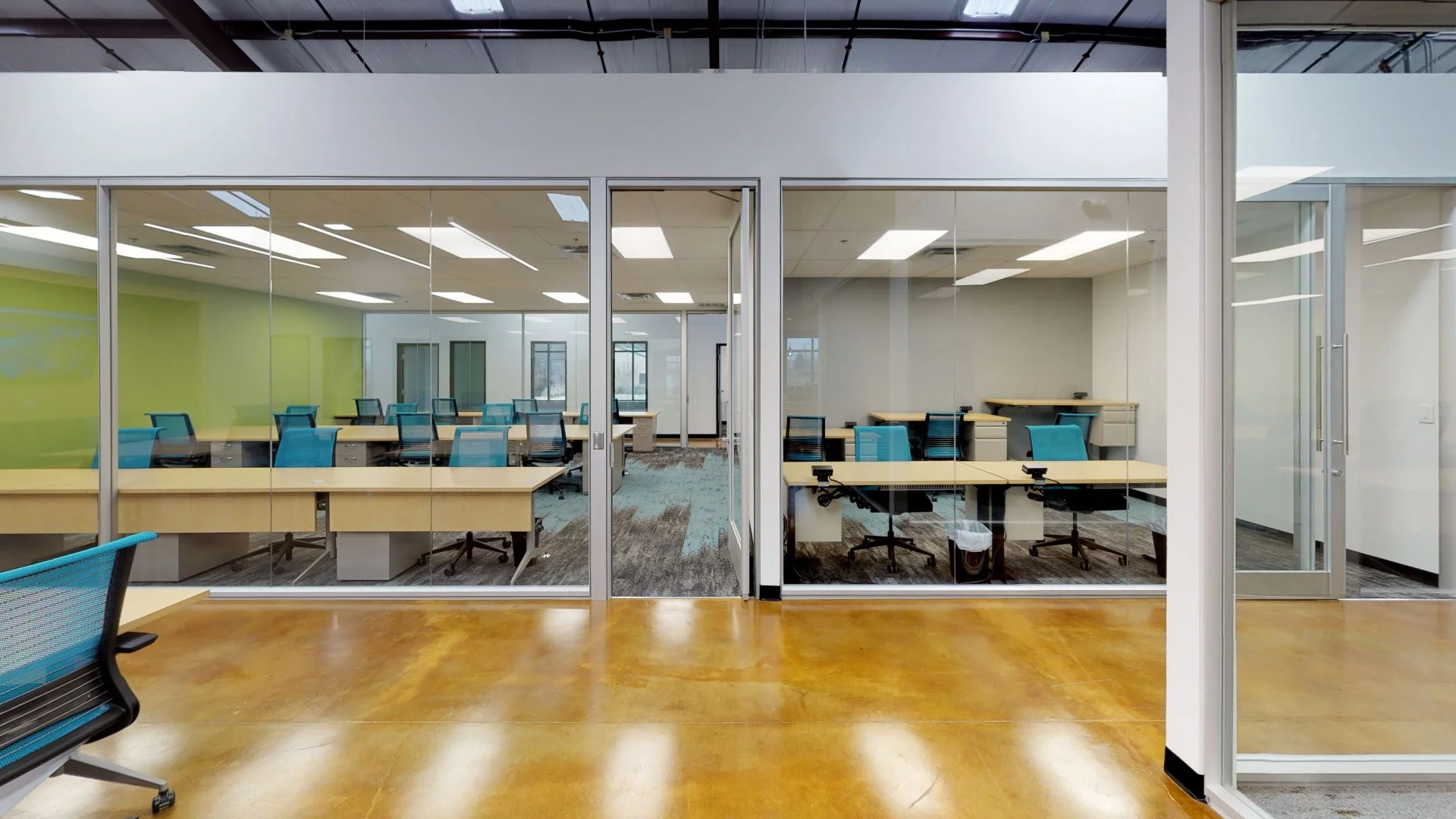 A modern, glass-enclosed training room featuring rows of light wood desks and bright blue ergonomic chairs, viewed from an open office hallway with polished amber-colored concrete floors.