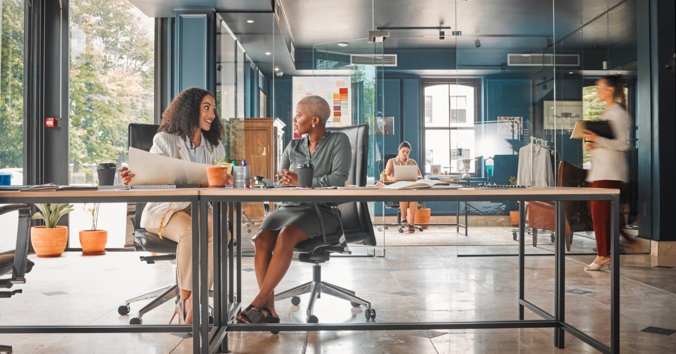 Two women engaged in conversation while sitting at a table in a modern office setting.